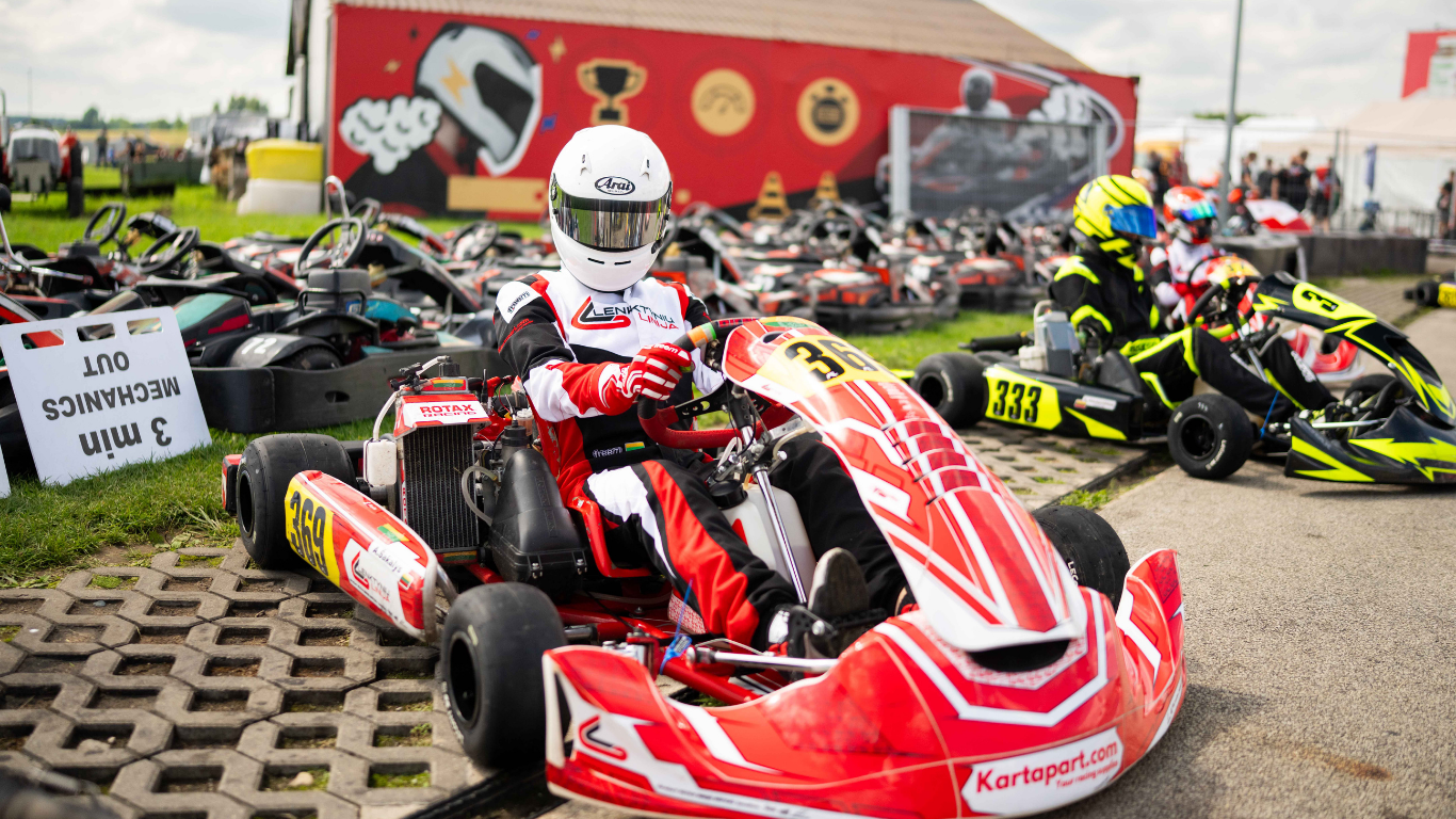 Kartingo varžybų dalyviai su lenktyniniais kombinezonais ir šalmais ruošiasi startui trasoje – dinamiška automobilių sportas akimirka prieš lenktynes.